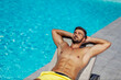 Sunbathing by the pool. A handsome man with muscles in his stomach is lying in a deck chair with his hands under his head and sunbathing. Self-tanning and enjoying by the pool on a sunny summer day
