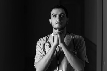 Wall Mural - Black and white portrait of praying male doctor on dark background