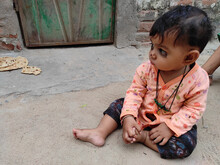 Close Up Of A Little South Asian Kid Standing On A Dirt Ground In Front Of A Rusty Wooden Door
