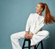 Smiling slim young woman with ponytail hairstyle in white trendy casual pantsuit, kimono style and fashionable sneakers sits on stool holding foot up and looks aside oat copy space
