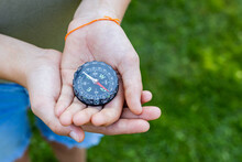 Girls Hands Holding The Compass. Hands Of A Teenager Girl Holding A Liquid Compass. Red Compass Needle Points North. Green Grass Background. Copy Space. Orienteering On The Ground. Direction Of Motion