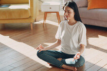 Asian Woman Doing Yoga And Zen Like Meditation In Lotus Pose In Casual Wear At Indoor Living Room Apartment With Natural Sun Light.