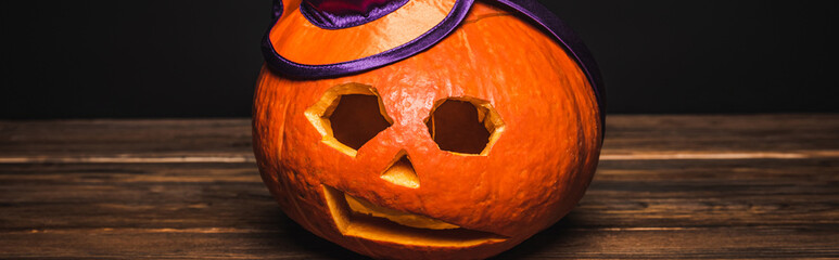 Poster - creepy and carved pumpkin in decorative hat on wooden surface isolated on black, banner