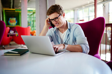 Successful male programmer in optical eyewear browsing content text while creating web graphic design on modern laptop computer, happy IT professional checking email messages while calling via mobile