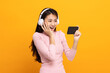 © Chanakon - Beautiful young asian women play mobile game and put on wireless headphone standing on isolated yellow background. Playing game on smartphone winning victory moment. Very enjoy and fun relax time