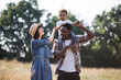 © sofiko14 - Beautiful multicultural family of three dressed in casual clothes walking together of summer field. African man holding boy in shoulders, caucasian woman embracing them.
