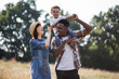 © sofiko14 - Little african boy sitting on father's shoulders and smiling sincerely to caucasian mother that walking near. Happy multiracial family outdoors. Summer time concept.