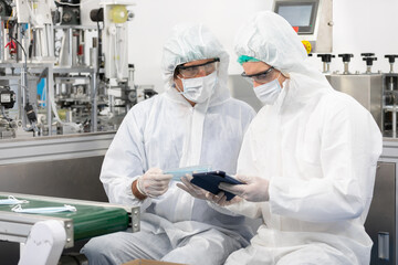 Wall Mural - male engineers wearing personal protective equipment uniform(PPE) using tablet and checking medical face mask beside machine in laboratory