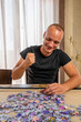 © Andres - Handsome man doing a puzzle alone and enjoying a relaxing day at home. Man in his 20s sitting on the chair with a board game on the table.