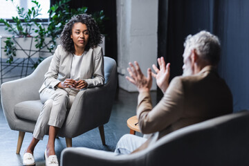 curly african american journalist looking at blurred businessman sitting in armchair and gesturing while talking during interview