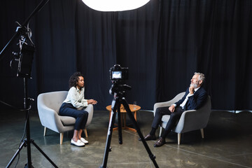 curly african american journalist sitting in armchair and talking with guest in studio