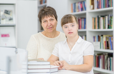 Sticker - Portrait of a senior woman and girl with syndrome down at library. Education for disabled children concept