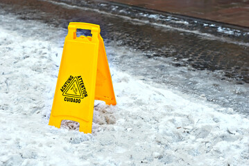 Yellow danger sign. Snow and ice on a slippery sidewalk. Inscription attention in different languages. Plate near the cobblestones in the spring.