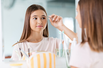 Poster - beauty, make up and cosmetics concept - teenage girl with tweezers tweezing her eyebrow and looking to mirror at home bathroom