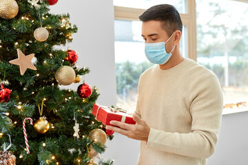 Wall Mural - winter holidays, health and people concept - middle aged man in medical mask with gift box at christmas tree at home