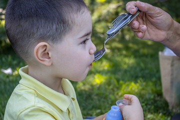 A close-up photo of a little kid (boy) dressed in a yellow t shirt  that is fed by his grandma with an iron spoon, in the park, and holds a plastic bottle in hands. Concept : ecology, healthy life.