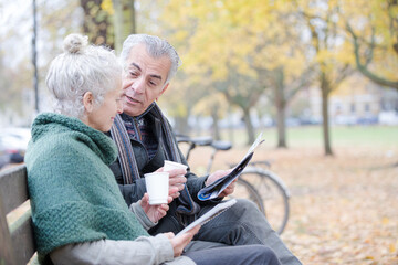 Wall Mural - Senior couple reading newspaper and drinking coffee on bench in autumn park