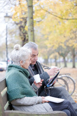 Wall Mural - Senior couple reading newspaper and drinking coffee on bench in autumn park