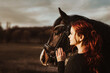 2 heads one love! stallion with head collar and redhead woman are bonding while being intimate friends and look left into sun to the future of cooperation of horse and rider enjoy the moment of trust