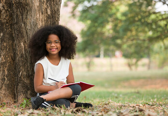 Wall Mural - Happy smiling African American little girl wearing glasses using pencil writing, drawing or doing homework on notebook under big tree. Children and education concept