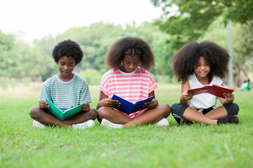 Wall Mural - Children reading book on grass outdoor. Group of African American children in casual wear reading book while sitting on green grass in the park, against green summer garden