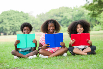 Wall Mural - Children reading book on grass outdoor. Group of African American children in casual wear reading book while sitting on green grass in the park, against green summer garden