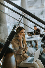 Wall Mural - Young woman sitting on the stairs with laptop and using mobile phone in modern office in front of her team