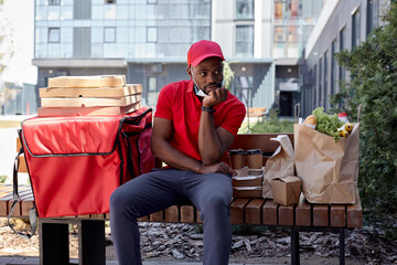 Wall Mural - Bored Afro american young Courier in red uniform and medical mask with thermal backpack delivering fresh lunches to clients, sit on bench waiting outdoors. Takeaway restaurant food delivery concept