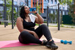 © Roman - Portrait Of Fat African Black Woman Sitting On Fitness Mat Having Rest Outdoors, Drinking Fresh Water, Enjoying Sport. Tired And Exhausted Female In Black Tracksuit Need Some Rest. Copy Space