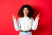 Attractive Caucasian Woman Showing Way, Pointing Fingers Sideways At Two Promos, Demonstrating Variants And Smiling, Standing On Red Background