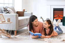 Young Mother With Daughter Reading Book Near Fireplace At Home