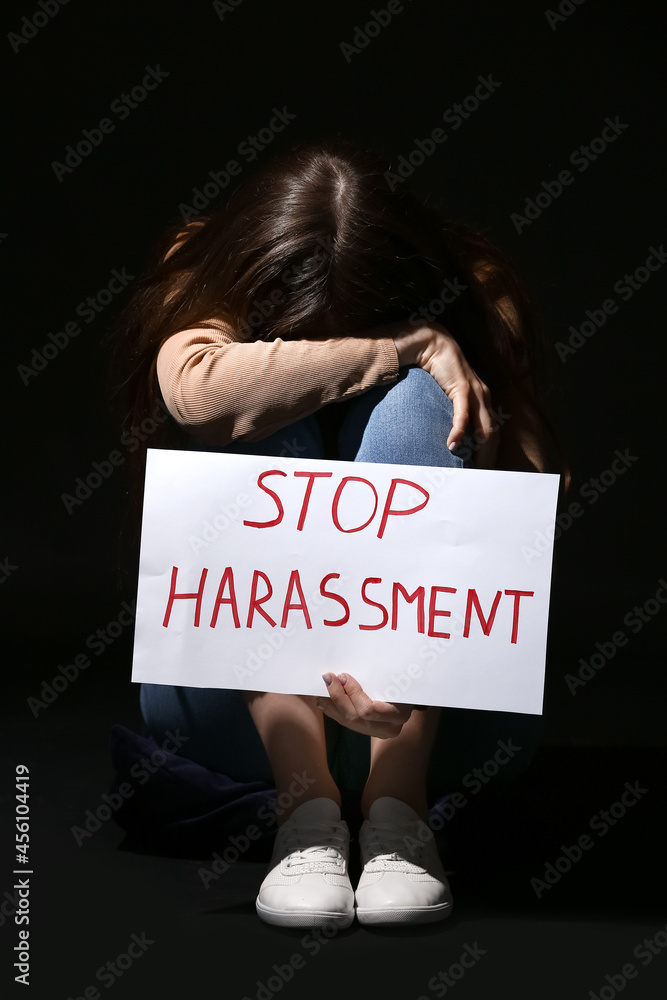 Stressed woman holding paper with text STOP HARASSMENT on dark background