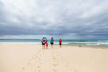 Teenagers And Children Walking Towards The Sea On Beach Under Stormy Clouds