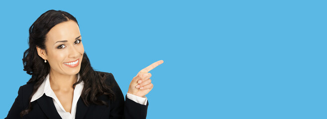 Sticker - Happy smiling attractive woman in black confident suit, pointing showing copy space area. Business concept. Bright blue colour background. Brunette businesswoman. Wide image.