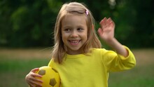 Portrait Of Little Girl With Yellow Soccer Ball Looks At Camera, Smiles And Laughs In Summer Park. Happy Child Waves Hand Outdoors. Football And Sport Concept.