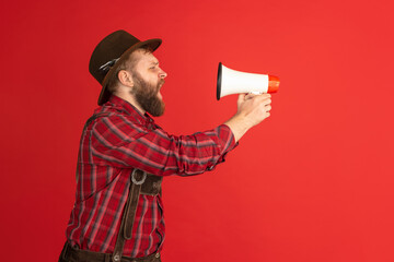 Wall Mural - Comic portrait of bearded man, waiter in hat and traditional Bavarian costume shouting at megaphone isolated over red studio background. Oktoberfest, fest, holidays concept