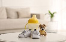 Baby bottle and shoes near toy bear on white table in room. Maternity leave concept
