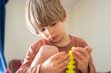 a kid plays with a toy at home. cute little five year old caucasian child boy with long blond hair playing hands with a yellow children's toy rubber caterpillar