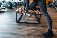Woman Athlete In Black Clothes With Dumbbells Doing Exercise On The Wooden Bench In The Gym