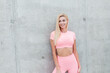 © alones - Pretty smiling fitness model woman with blond hair in fashion pink sportswear outfit near gray concrete wall in the city