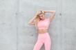 © alones - portrait of young fitness model woman with blonde hair in fashionable pink sportswear posing near gray concrete wall in the city