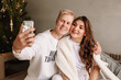 © Елизавета Старкова - Happy man and woman in love in cozy sweaters sitting on the couch taking selfies communicating by video and congratulating on the Christmas holiday online using a mobile phone at home, selective focus