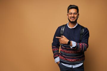 Wall Mural - Smiling happy indian student with backpack pointing his finger on background