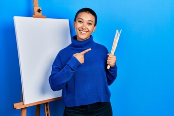 Sticker - Beautiful hispanic woman with short hair holding brushes close to easel stand smiling happy pointing with hand and finger