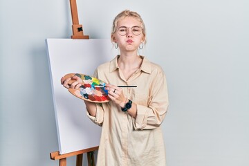 Wall Mural - Beautiful caucasian woman with blond hair standing drawing with palette by painter easel stand puffing cheeks with funny face. mouth inflated with air, catching air.