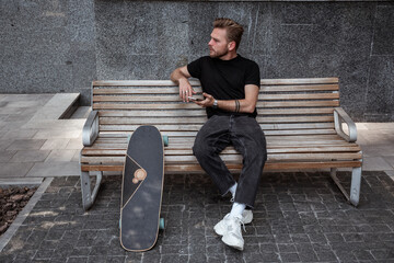 Serious young man with longboard sitting on bench in modern city location looking at the distance holding smartphone, surfing internet, reading news, liking posts. Outdoor leisure concept.