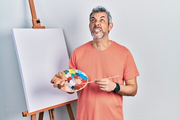 Sticker - Handsome middle age man with grey hair standing drawing with palette by painter easel stand angry and mad screaming frustrated and furious, shouting with anger looking up.