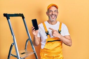 Sticker - Handsome middle age man with grey hair standing by ladder showing smartphone smiling and laughing hard out loud because funny crazy joke.