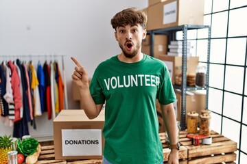 Poster - Young arab man wearing volunteer t shirt at donations stand surprised pointing with finger to the side, open mouth amazed expression.