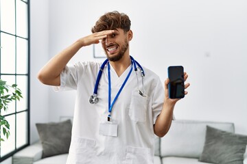 Sticker - Young arab man wearing doctor uniform and stethoscope holding smartphone stressed and frustrated with hand on head, surprised and angry face